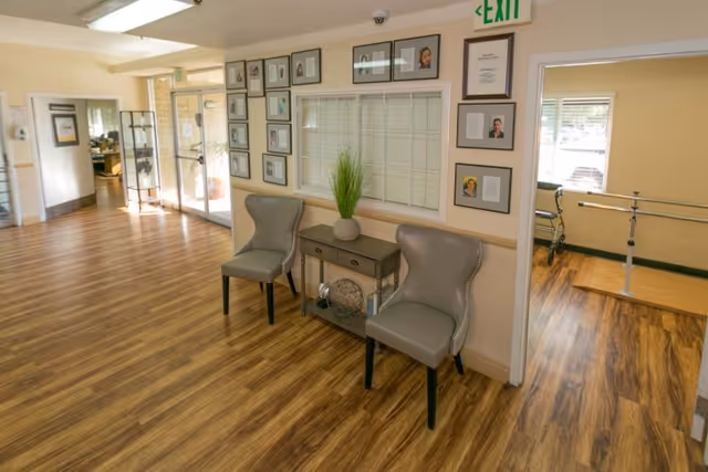 Interior view of a senior living facility hallway with wood flooring, two gray chairs flanking a small table with a plant, framed photos and certificates on the walls, a window with blinds, and an exit sign above a doorway leading to a room with parallel bars for physical therapy.