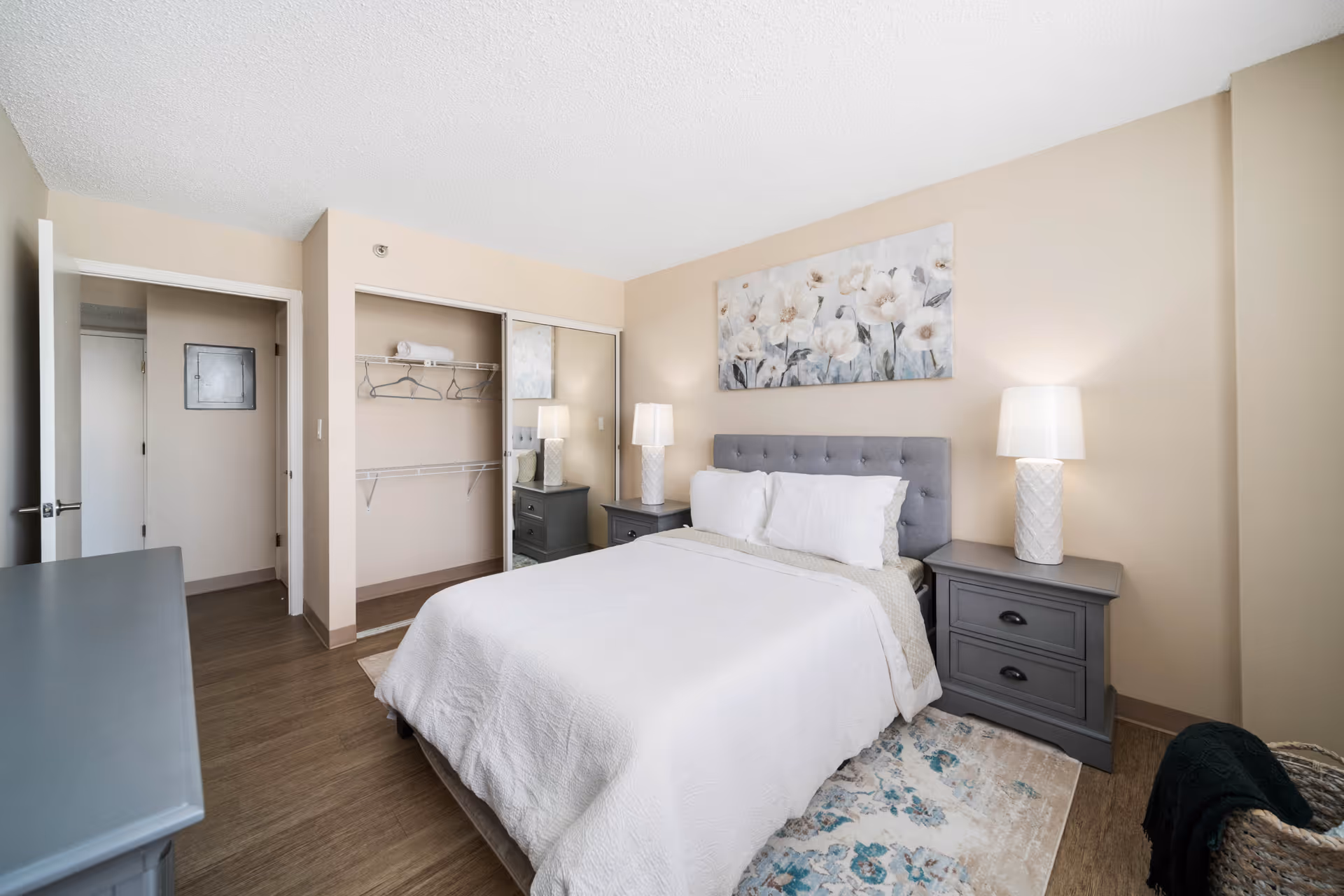 A neatly arranged bedroom with a queen-sized bed covered in white bedding. The bed has a gray tufted headboard and is flanked by two gray nightstands, each with a white lamp. Above the bed hangs a large floral painting. There is an open closet with sliding mirrored doors, a few hangers, and a rolled towel on the shelf. The room has beige walls, wood flooring, and a patterned area rug partially under the bed. A woven basket with a dark blanket is visible in the corner.
