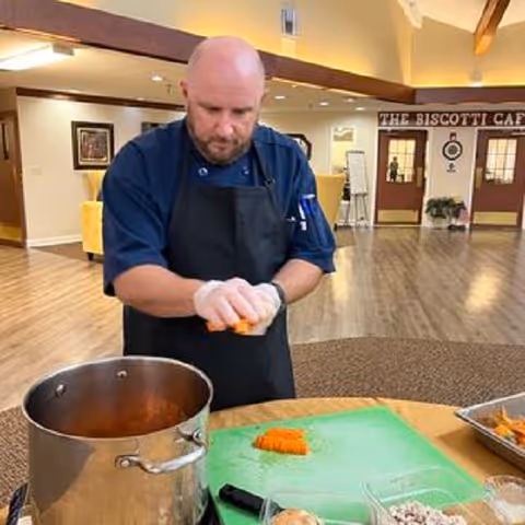 A man wearing a black apron and gloves is peeling or cutting carrots at a round table with a large pot and other food items in front of him. The background shows a spacious room with wooden floors, yellow chairs, and a sign that reads 'THE BISCOTTI CAFE' above two doors.
