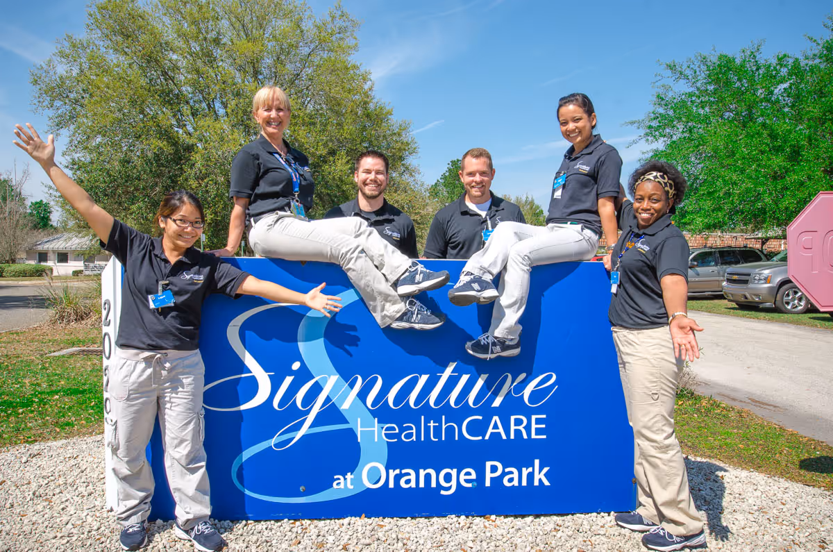 Six staff members in matching uniforms pose and smile around a large blue 'Signature HealthCARE at Orange Park' outdoor sign.