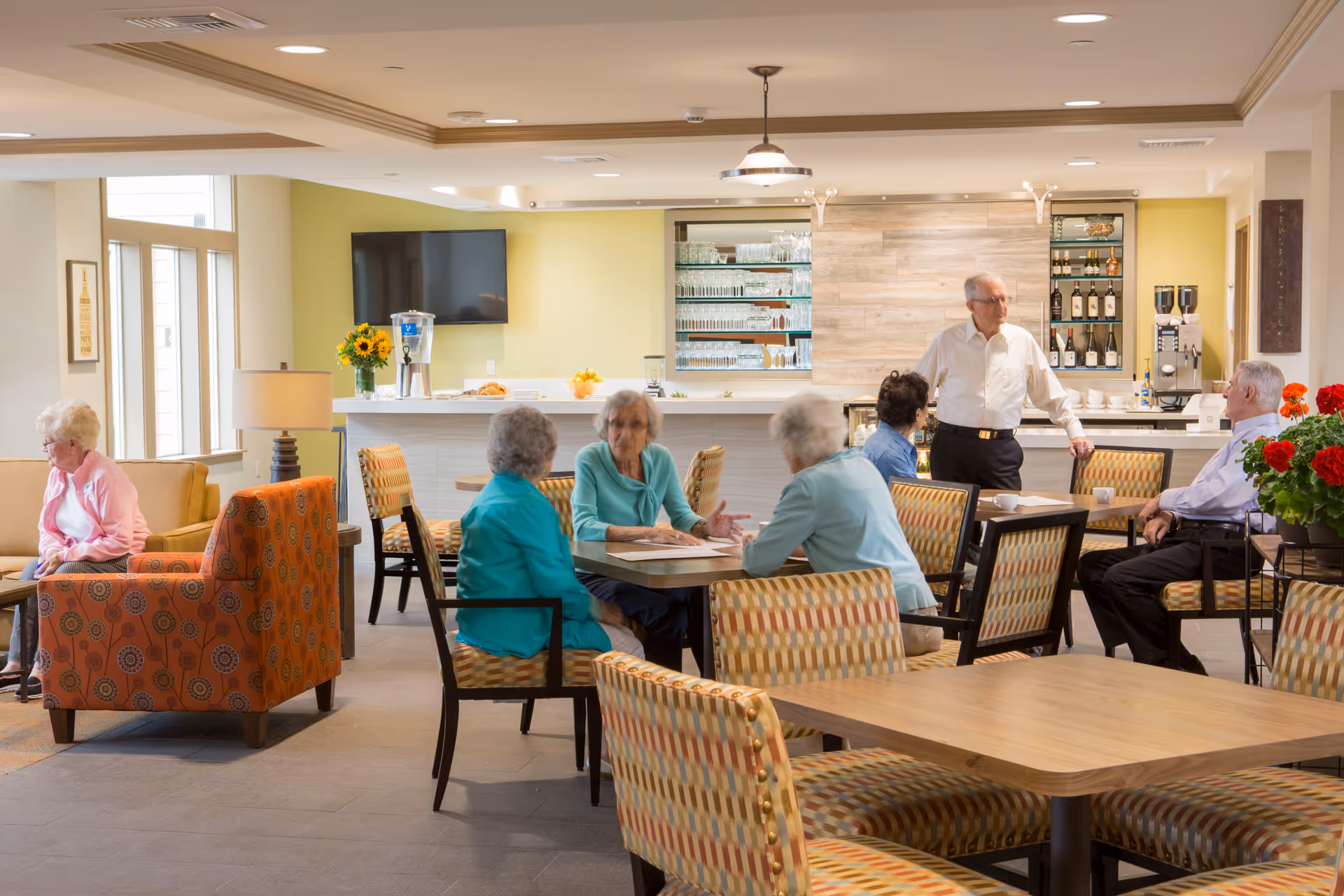 Several older adults seated and talking at tables in a bright communal dining and lounge area with a service counter and patterned chairs.