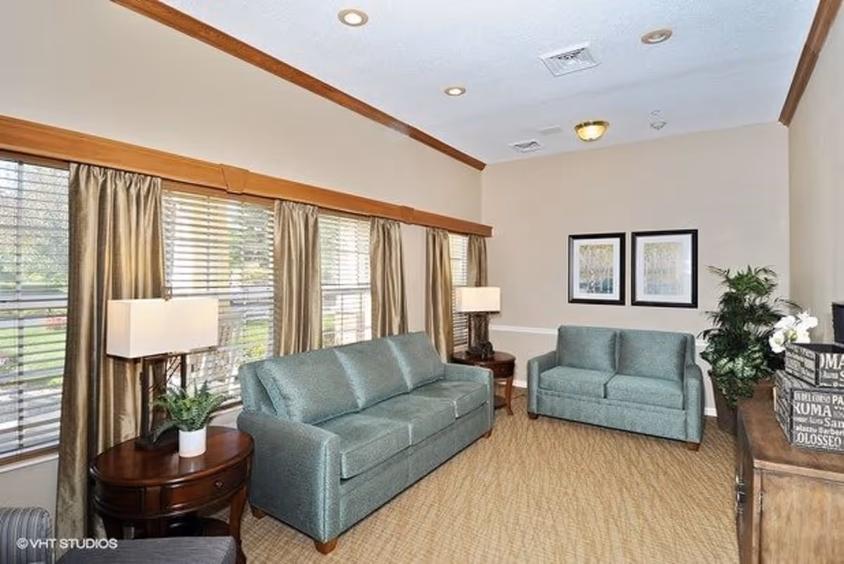 A cozy living room with two teal sofas, a wooden side table with a lamp and a small plant, beige walls with two framed pictures, a wooden cabinet with decorative boxes, a potted plant, and large windows with beige curtains allowing natural light to enter.