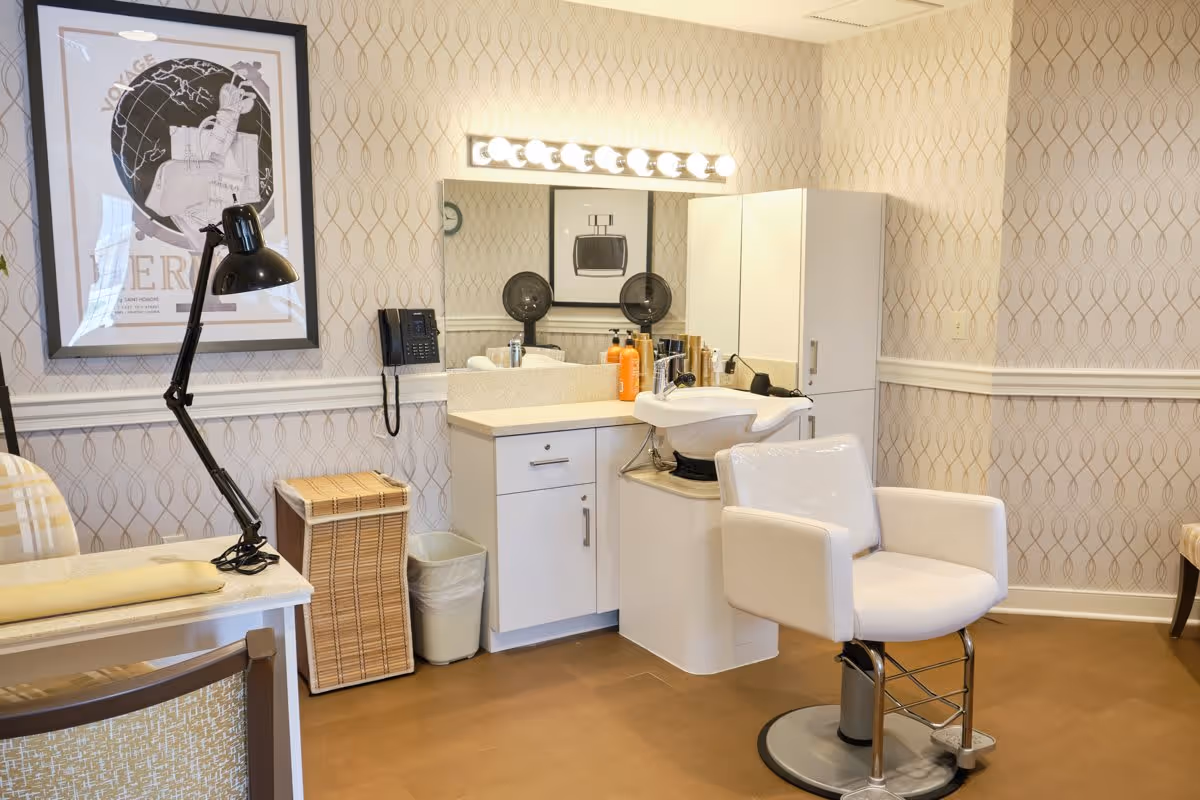 A well-lit salon area inside a senior living facility featuring a white salon chair in front of a sink and countertop with hair care products. The wall has patterned wallpaper and a large mirror with lights above it. There is a black adjustable desk lamp on a table with a plaid chair, a wicker laundry basket, a trash bin, and framed artwork on the walls.