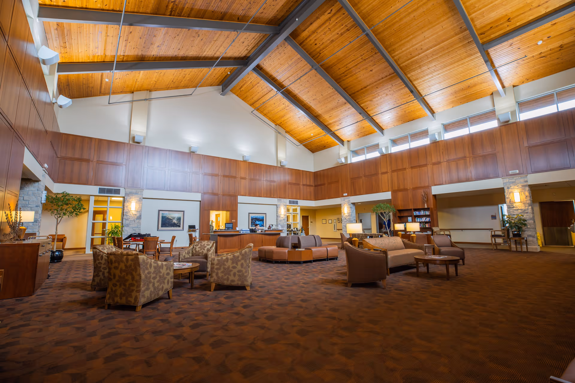 Spacious senior living facility common area with high wooden ceiling, large windows near the ceiling, wood-paneled walls, and warm lighting. The room is furnished with multiple seating arrangements including armchairs, sofas, and small tables. There is a reception desk in the background and some indoor plants and framed pictures on the walls.