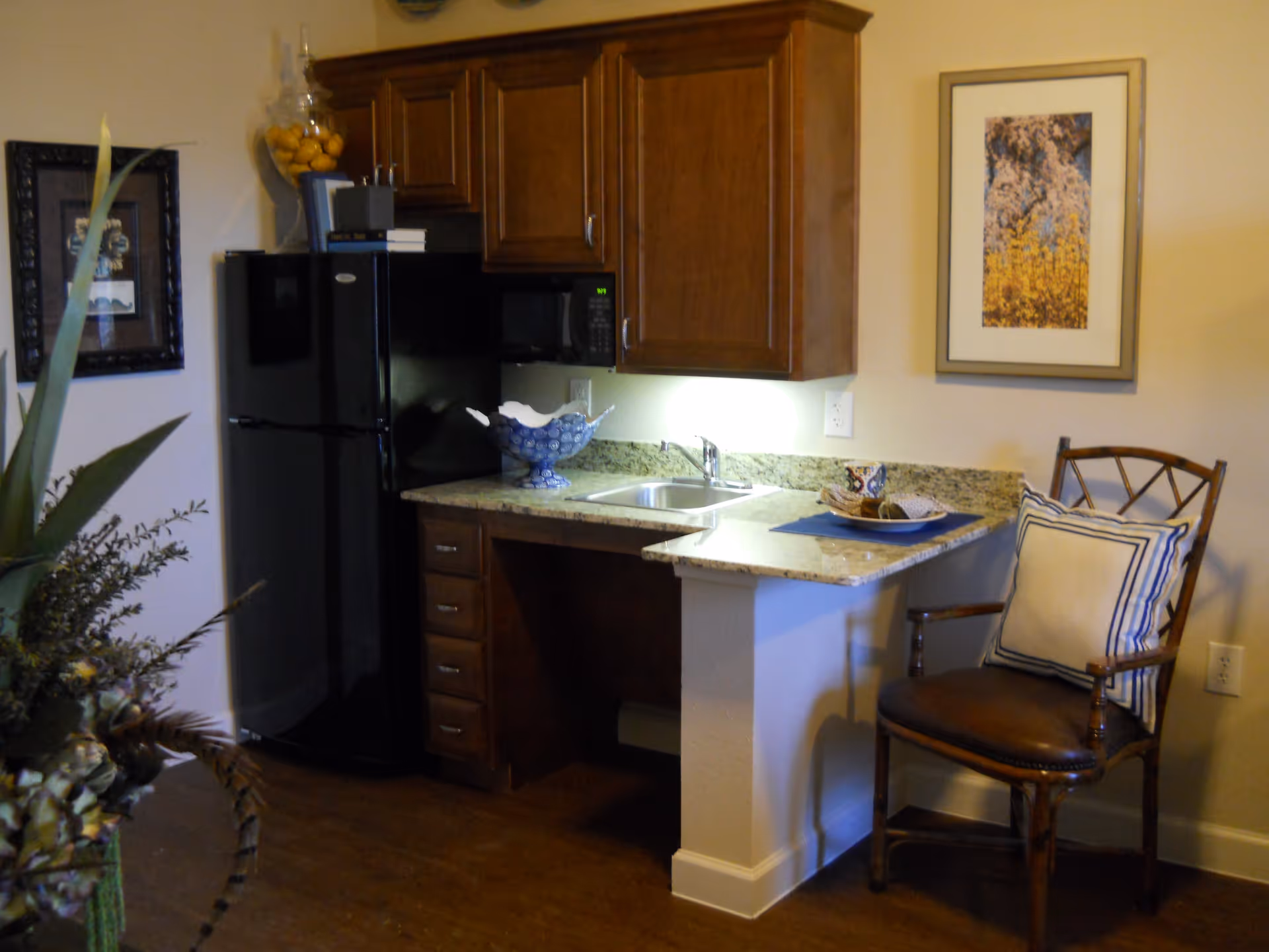 A small kitchen area with wooden cabinets, a black refrigerator, a microwave, a sink, and a granite countertop. There is a decorative bowl on the counter and a wooden chair with a cushion next to the counter. The walls have framed artwork and the floor is wooden.