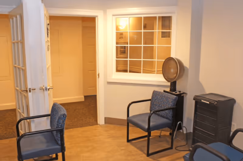 A small waiting area with three blue cushioned chairs with black metal frames, a vintage hair dryer chair, and a black rolling cart. The room has beige walls, a window with white trim, and an open door leading to a carpeted hallway.
