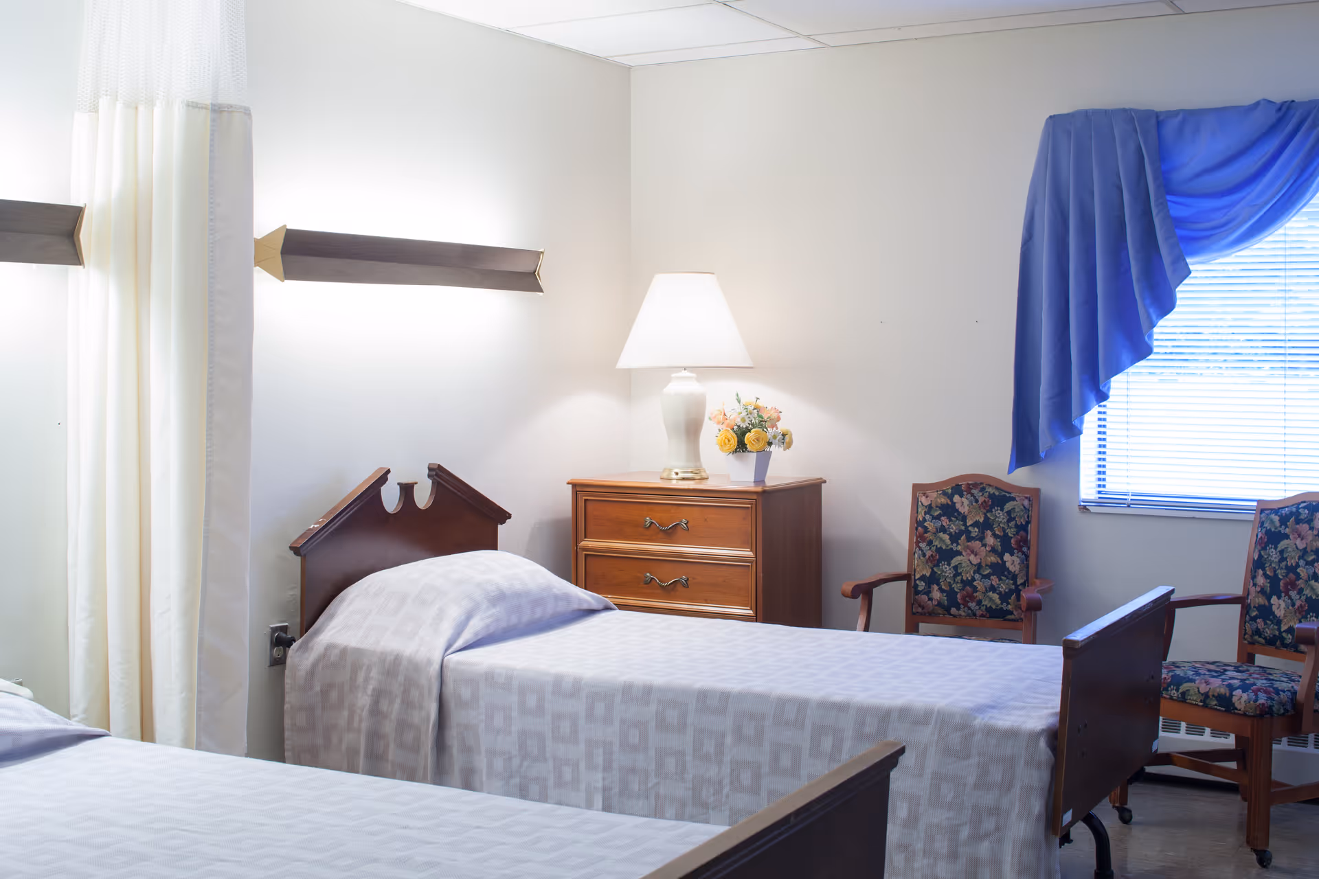 Twin-bed senior care room with a nightstand and lamp, floral chairs, and a curtained window.