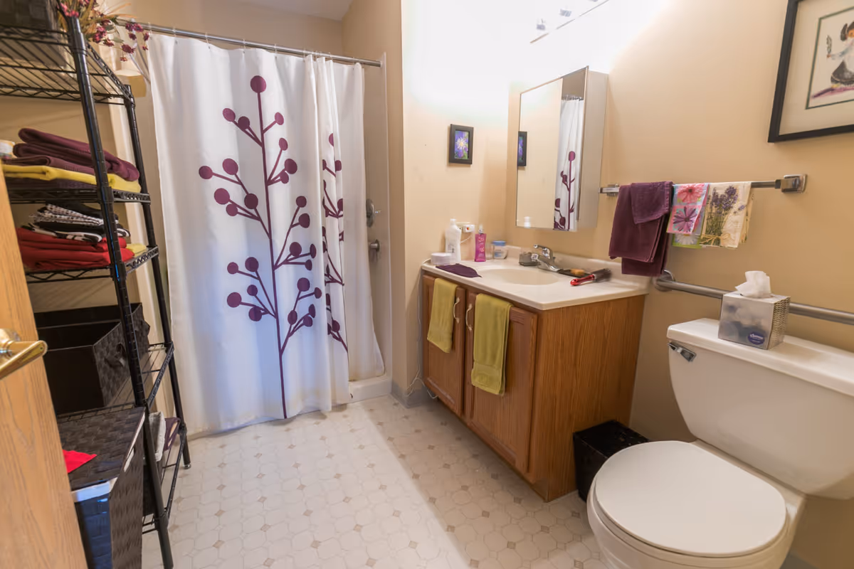 A bathroom with a white shower curtain featuring a purple branch design, a wooden vanity with a white countertop and sink, a mirror above the sink, and a toilet with a tissue box on top. There are towels hanging on a rack and a metal shelving unit with folded towels and baskets on the left side.