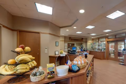 Interior view of a facility's reception or common area with a counter displaying baskets of fruit, snacks, and other items. The area is well-lit with ceiling lights and has wooden cabinetry and glass display cases in the background.