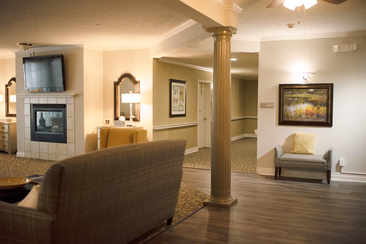 A cozy living room area in a senior living facility featuring a fireplace with a mounted TV above it, a beige armchair with a lamp and mirror behind it, a column in the center, a small bench with a yellow pillow, and framed artwork on the walls. The flooring is a combination of carpet and wood.