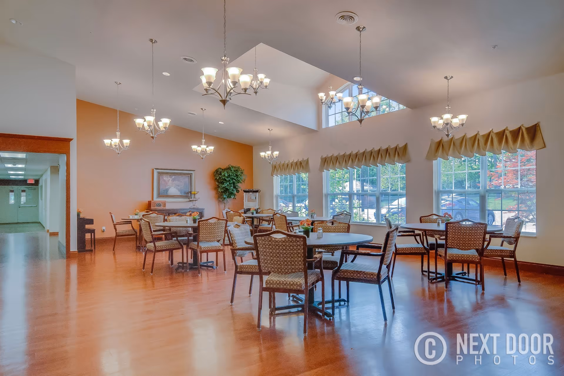 A bright dining room with multiple round tables and chairs arranged neatly. The room features large windows with beige valances, allowing natural light to fill the space. Several chandeliers hang from the ceiling, and there is a painting and a potted plant against a peach-colored wall. The floor is wooden, and a hallway is visible to the left.