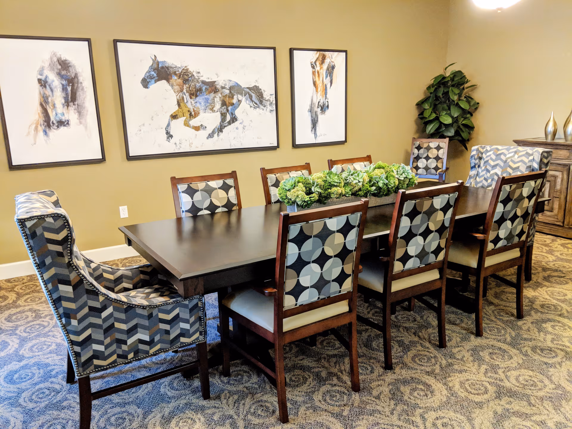 A dining room with a long dark wooden table surrounded by eight chairs, six with patterned backs and two larger armchairs with a chevron pattern. The room has beige walls adorned with three framed horse paintings. A green leafy plant is placed in the corner near a wooden sideboard with decorative items on top. The carpet has a circular pattern in neutral tones.