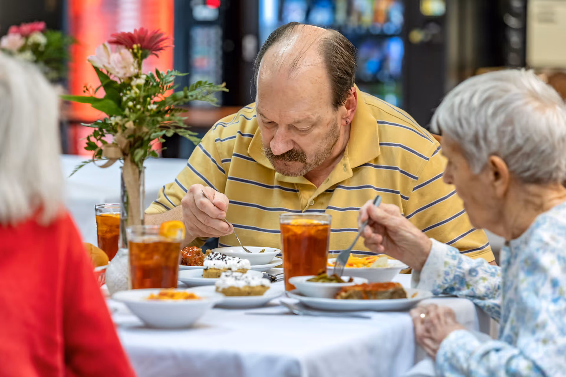 Three elderly people sitting at a table in a dining area, eating a meal with plates of food and glasses of iced tea. A vase with flowers is on the table.