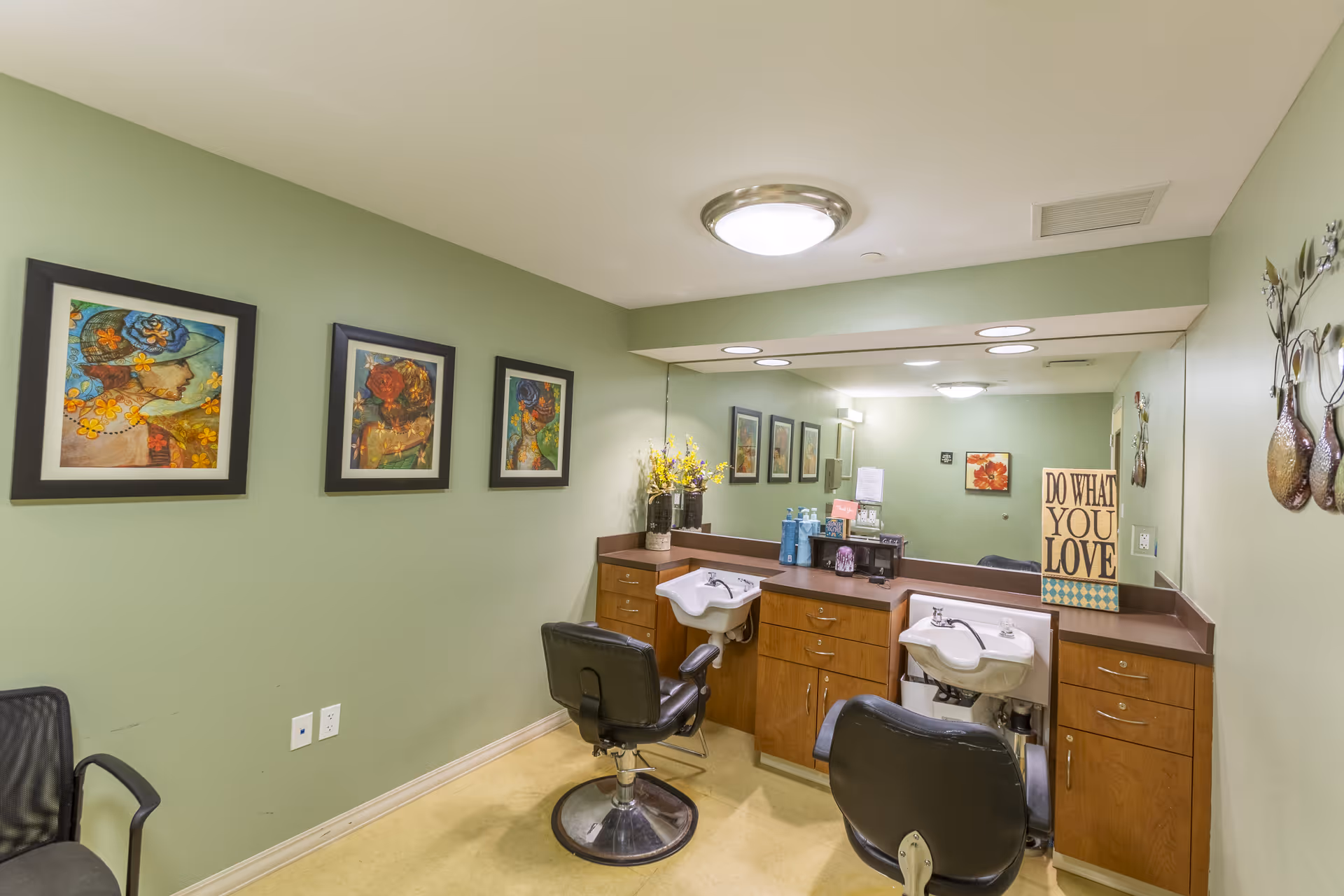 Interior view of a salon area with two black salon chairs in front of sinks and wooden cabinetry. The walls are painted light green and decorated with framed artwork and a sign that reads 'DO WHAT YOU LOVE'. A large mirror spans the wall above the sinks and cabinets.