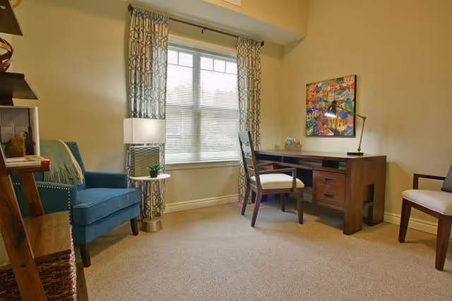 A bright sitting room with a wooden desk and chairs, a blue armchair by a window with patterned curtains, and a floor lamp.