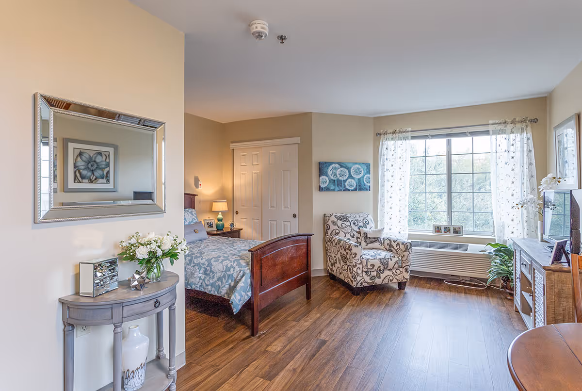 A bright and cozy senior living bedroom with a wooden bed covered in blue floral bedding, a patterned armchair near a large window with sheer curtains, a wooden dresser with a TV, and a small round table with a vase of flowers and decorative items. The room has wooden flooring and beige walls.
