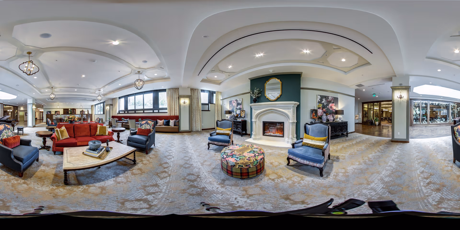 Spacious and elegant living room area in a senior living facility with comfortable seating including red and blue armchairs and sofas, a fireplace with a decorative mirror above it, large windows allowing natural light, and tasteful decor including floral paintings and a colorful round ottoman.