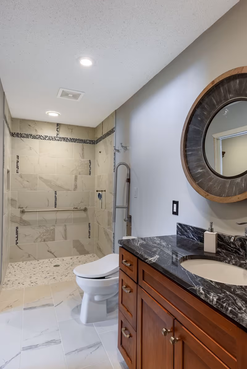 A modern bathroom featuring a walk-in shower with beige and black tiled walls, a white toilet, and a wooden vanity with a black marble countertop and an oval sink. A round mirror with a wooden frame hangs above the vanity.