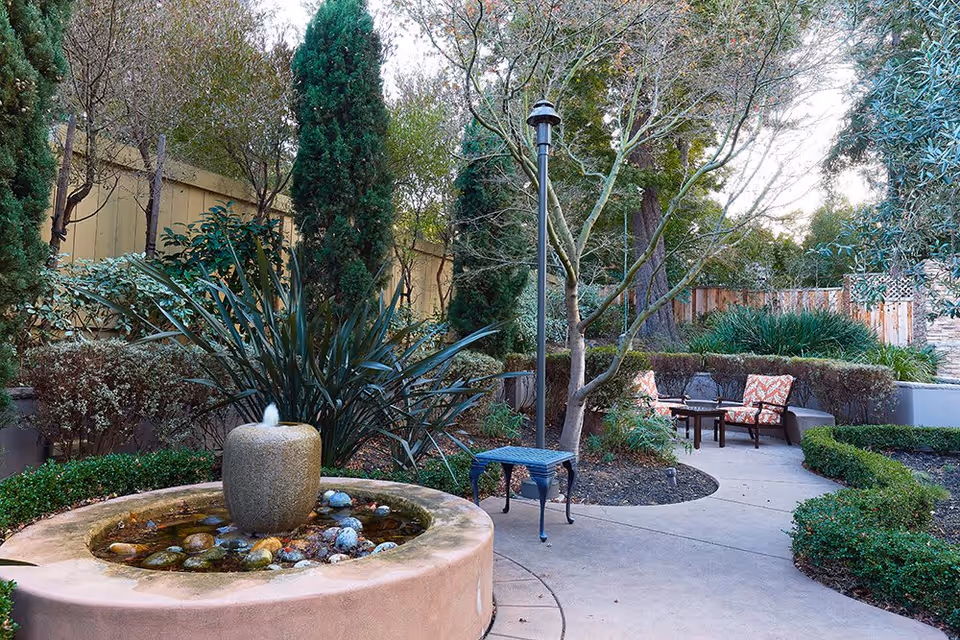 Outdoor garden area with a circular stone water fountain surrounded by plants and trees. There is a curved concrete pathway leading to a seating area with cushioned chairs and a small table. The area is enclosed by a wooden fence and has a lamppost near the pathway.