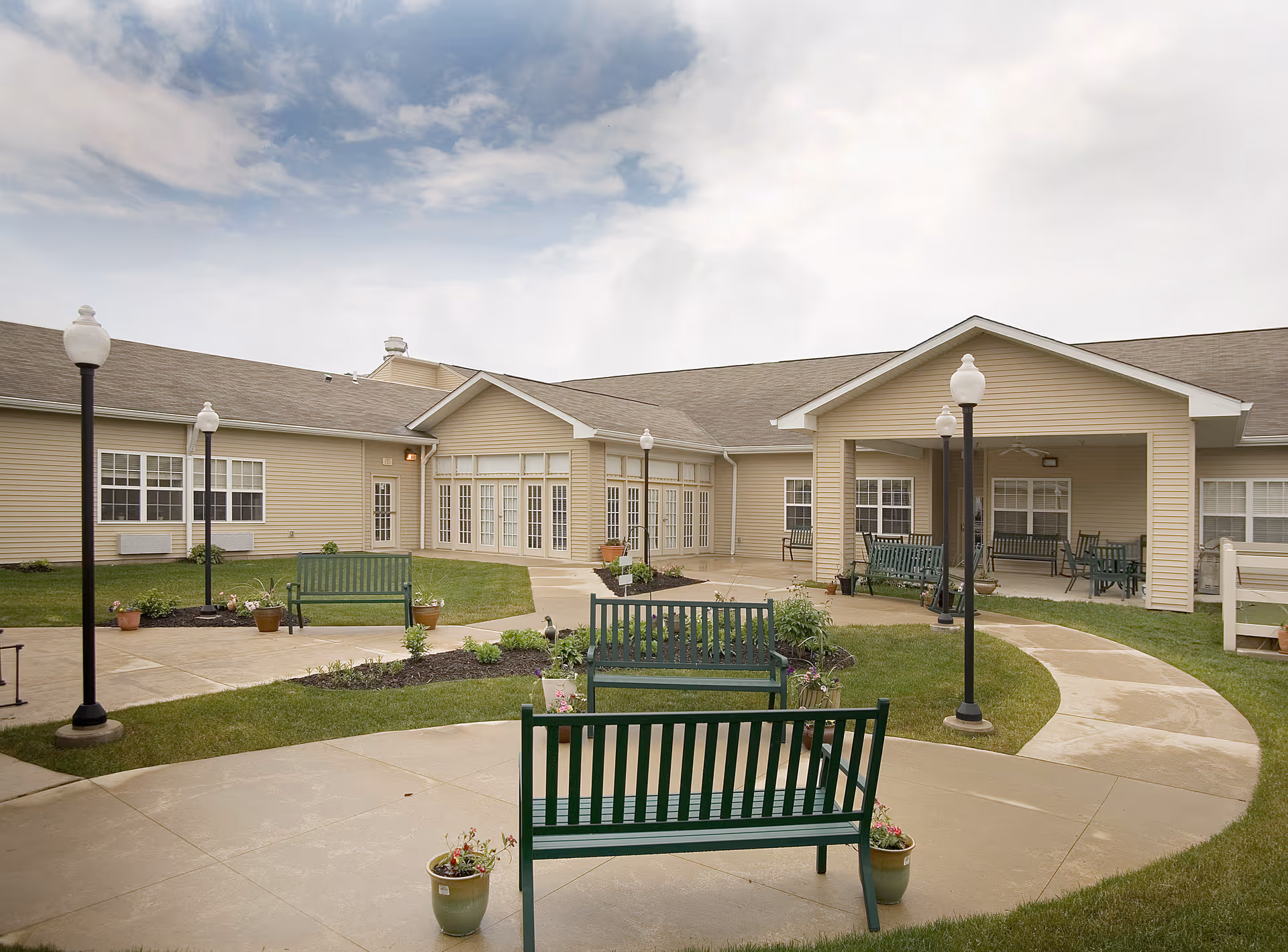 Outdoor courtyard area of a senior living facility with green benches, potted plants, lamp posts, and a paved walkway surrounded by grass. The beige building with multiple windows and a covered patio area is visible in the background under a partly cloudy sky.
