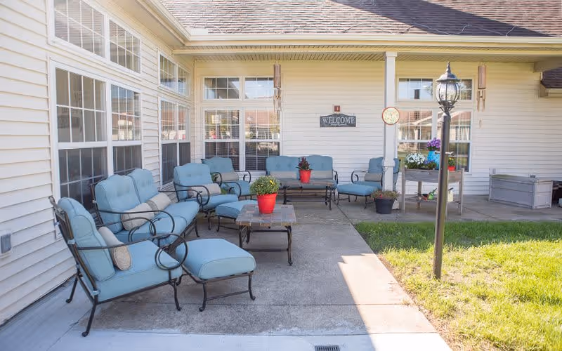 Outdoor patio area with cushioned metal chairs and sofas arranged around a coffee table with potted plants. The patio is adjacent to a building with large windows and a welcome sign on the wall. There is a lamp post on the grass next to the patio.