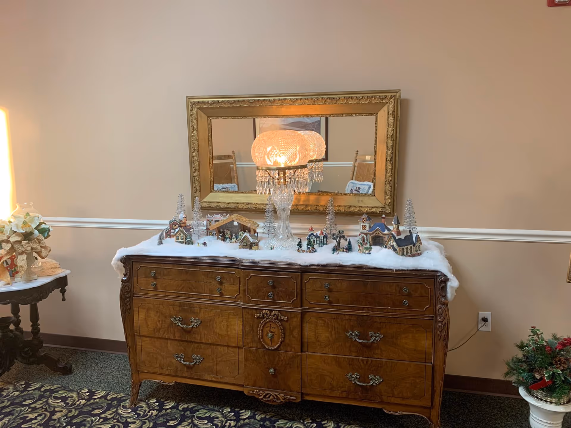 Ornate wooden dresser topped with a miniature snowy village display and a decorative lamp beneath a framed mirror in an interior room.