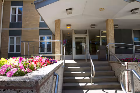 Entrance to a building with stairs and handrails leading up to glass doors. Flower beds with pink and purple flowers are on either side of the stairs. The building exterior is made of brick and concrete.