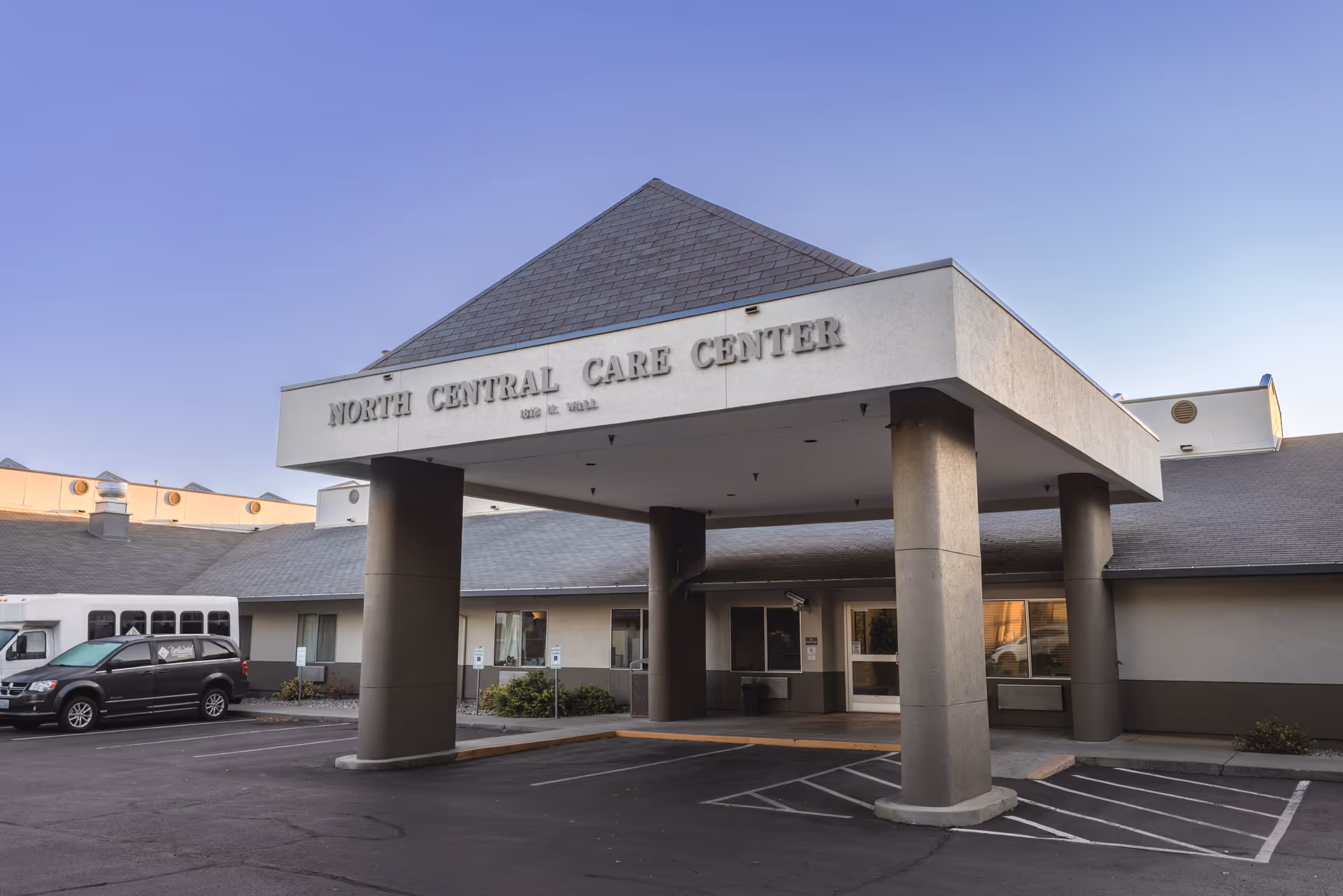 Exterior front entrance of North Central Care Center, showing a covered drop-off area supported by large columns, with a parking lot in front and a clear sky above.