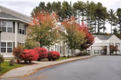 Exterior view of a senior living community building with beige siding and white trim. The building is surrounded by trees with autumn foliage in shades of red, orange, and green. A paved driveway and sidewalk lead to the entrance of the building.