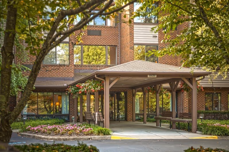 Entrance of a senior living facility named The Park Oak Grove, featuring a covered drop-off area with benches, surrounded by trees and flower beds, with a brick building in the background.