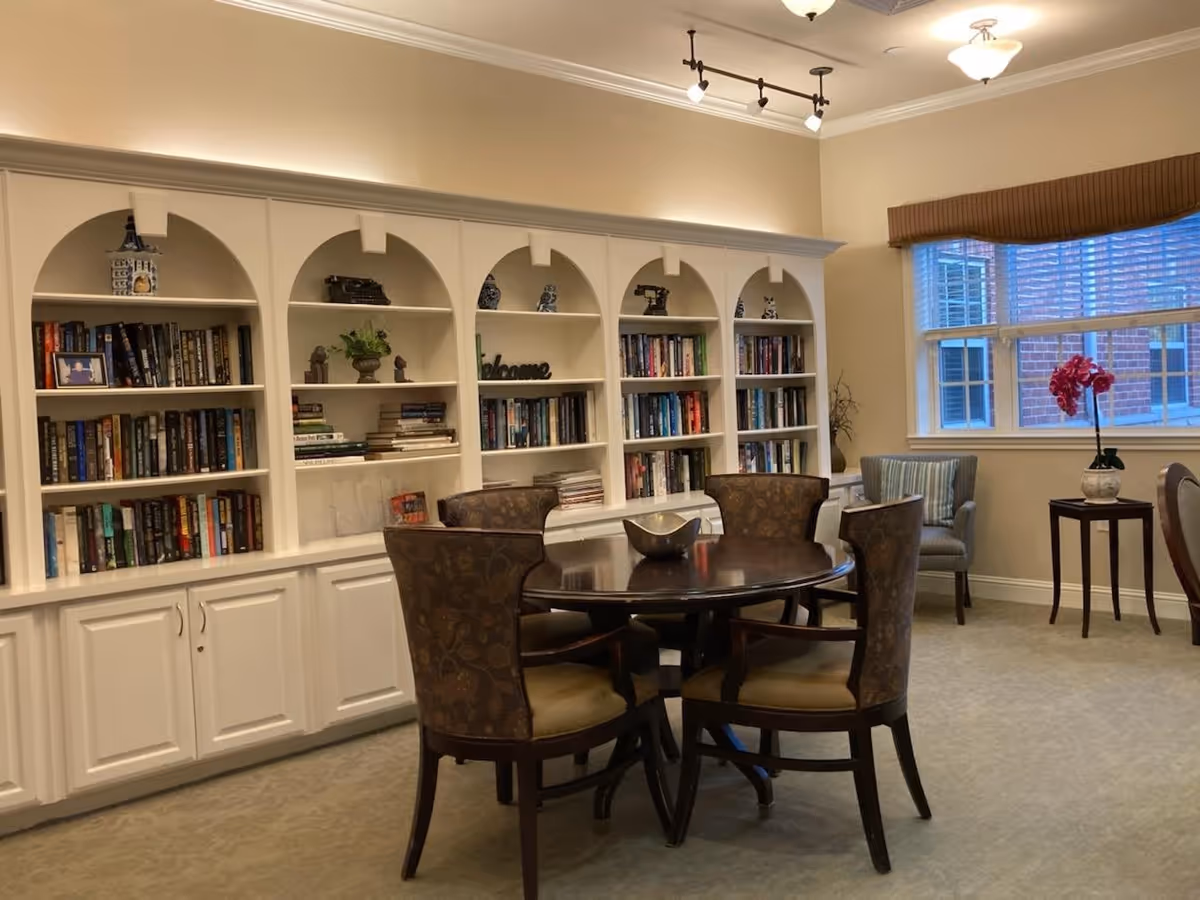 A cozy interior room with a round wooden table surrounded by four upholstered chairs. Behind the table is a large built-in white bookshelf filled with books and decorative items. To the right, there is a window with blinds and a small side table holding a potted plant with red flowers. The room is softly lit with ceiling lights.