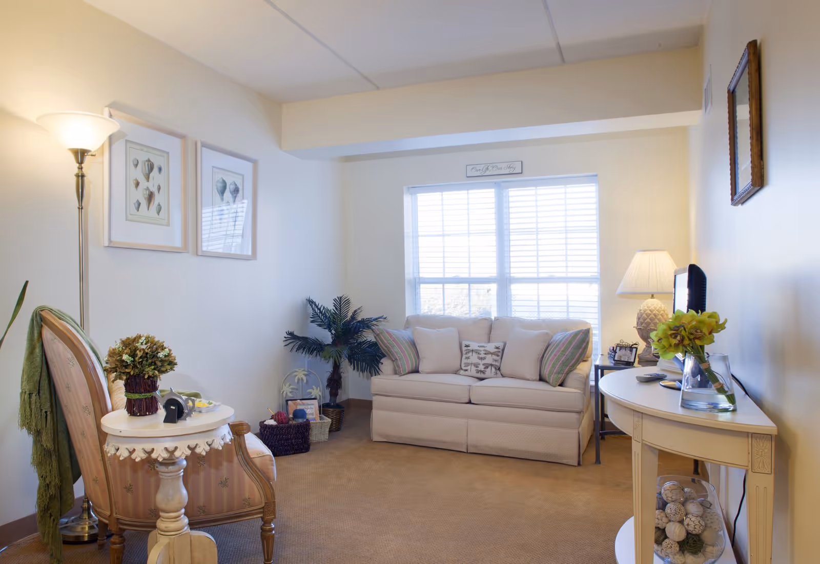 A cozy living room with a beige sofa adorned with multiple pillows, a wooden armchair with a green throw draped over it, a small round side table with a flower arrangement, a floor lamp, and a console table with a vase of flowers and decorative items. The room has a large window with white blinds letting in natural light, and framed artwork on the walls.