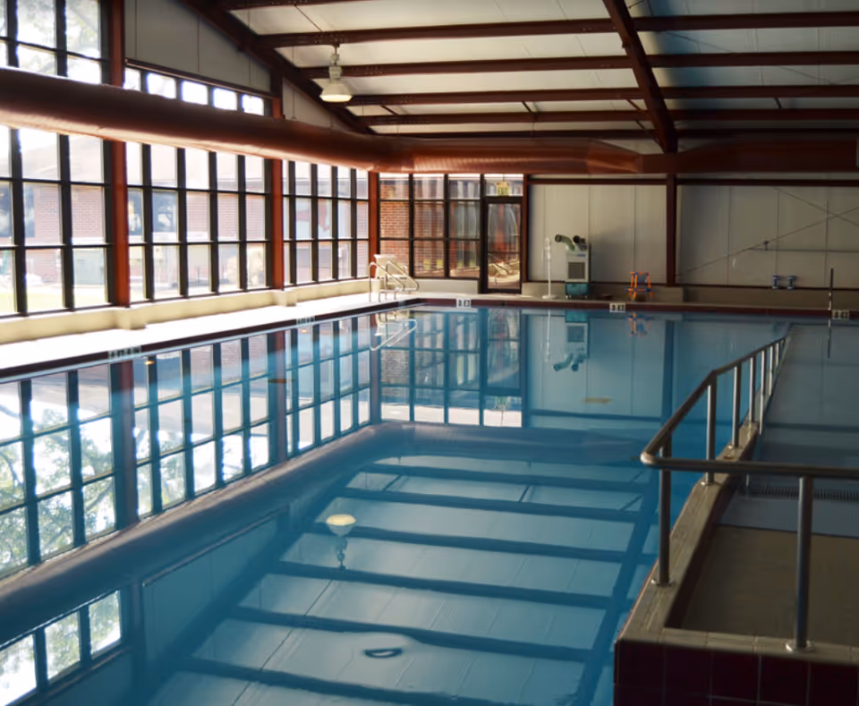 Indoor swimming pool with clear blue water, large windows along one side letting in natural light, metal handrails, and a ceiling with exposed beams and ventilation ducts.