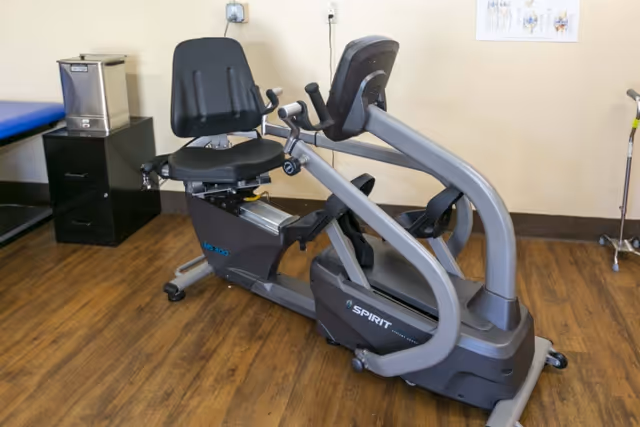 A recumbent exercise bike with a black seat and handles, placed on a wooden floor in a room with beige walls. There is a black cabinet with a metal container on top and a blue padded table to the left. A medical chart is partially visible on the wall in the background.
