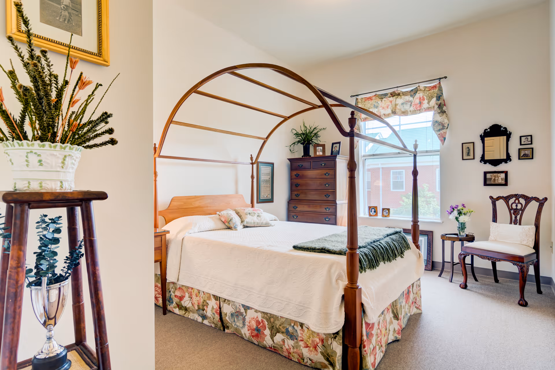 A bright bedroom with a wooden four-poster bed covered in white bedding and floral bed skirt. There is a wooden chest of drawers against the wall, a window with floral valance, a wooden chair with a cushion, and a small side table with a vase of flowers. A plant in a white pot sits on a wooden stand in the foreground.
