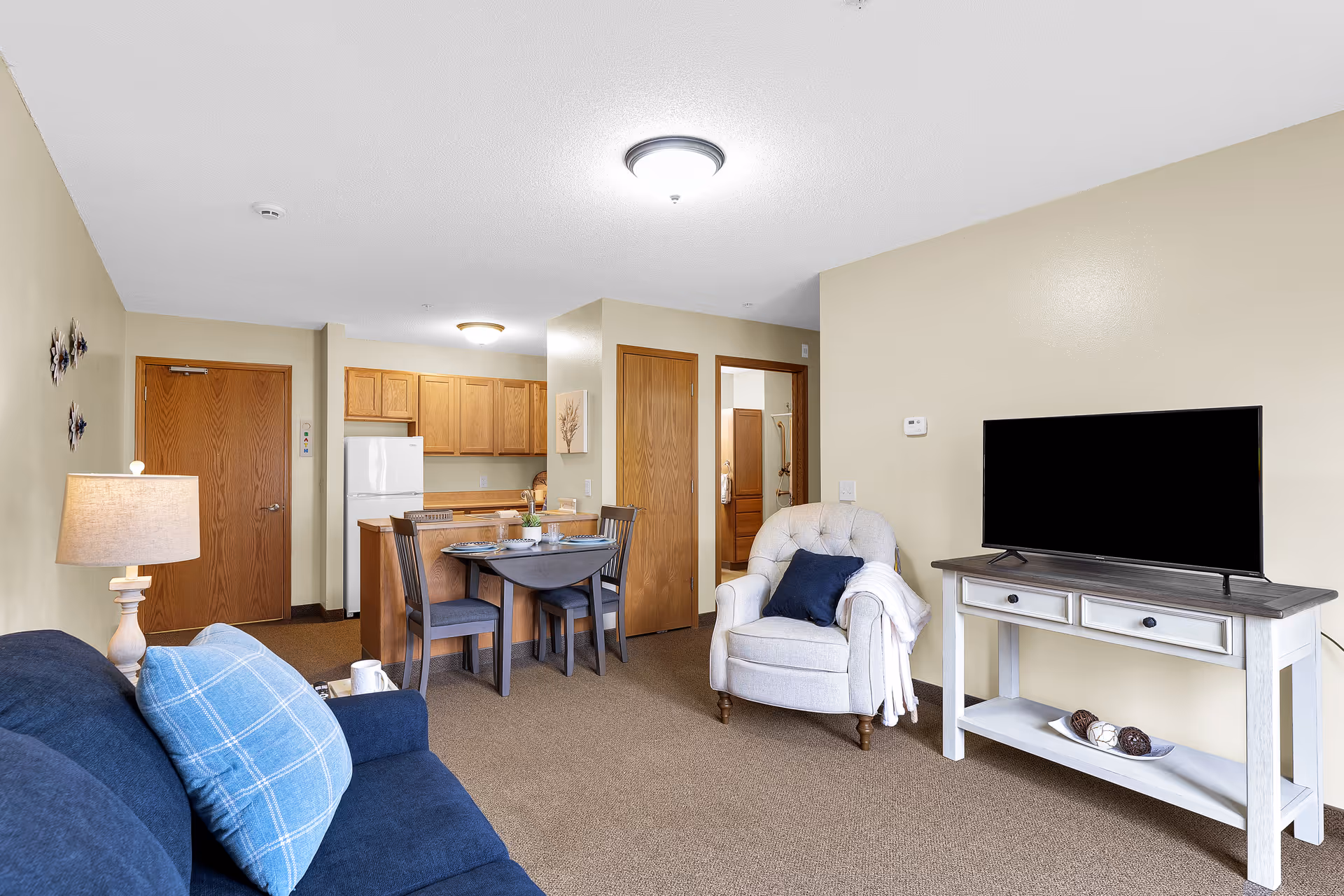 A cozy living area in a supportive living facility featuring a navy blue sofa with a blue plaid pillow, a beige armchair with a navy pillow and white throw blanket, a white TV stand with a flat-screen TV, and a small dining table with four chairs. The background shows a kitchen area with wooden cabinets, a white refrigerator, and two wooden doors leading to other rooms.