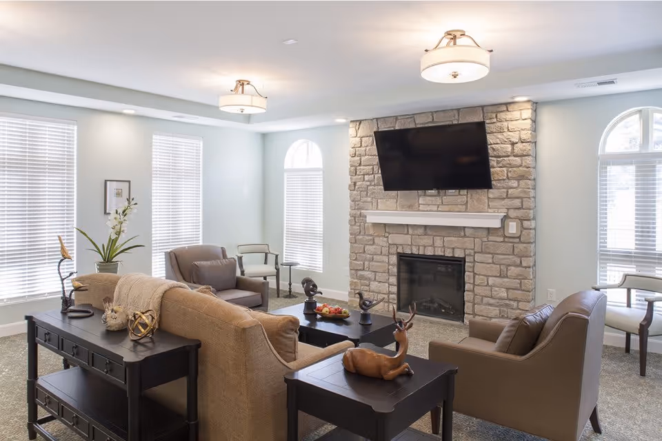 A cozy living room with a stone fireplace and a mounted flat-screen TV above it. The room features a beige sofa, two brown armchairs, a coffee table with decorative items, and side tables with ornaments. Large windows with white blinds allow natural light to fill the space, and the walls are painted a light color. Ceiling lights provide additional illumination.