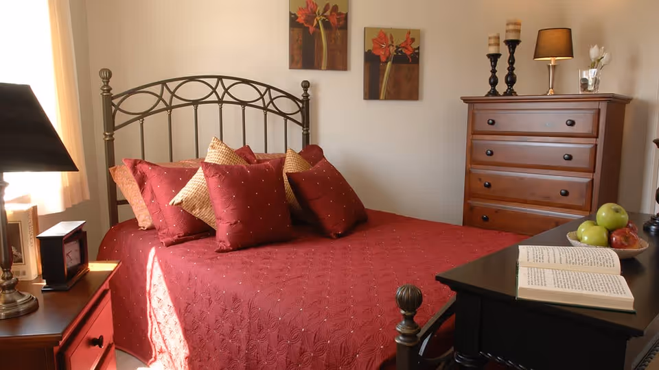 A cozy bedroom with a metal frame bed covered in a red quilt and multiple red and beige pillows. There is a wooden nightstand with a lamp and a clock on the left side of the bed. On the right side, there is a black table with an open book and a bowl of green apples and a red apple. A wooden dresser with candles and a lamp is against the wall, and two floral paintings hang above the bed.