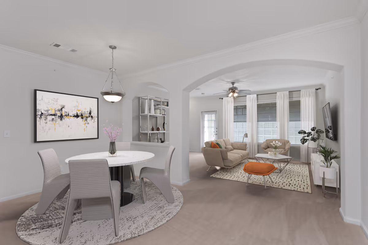 A bright and modern open living and dining area in an active adult apartment. The dining area features a round white table with four gray chairs on a patterned rug, and a vase with pink flowers as a centerpiece. The living room has a beige sofa, an armchair, an orange ottoman, a coffee table, and a TV mounted on the wall. Large windows with white curtains allow natural light to fill the space, and there are plants and decorative items on shelves and stands.
