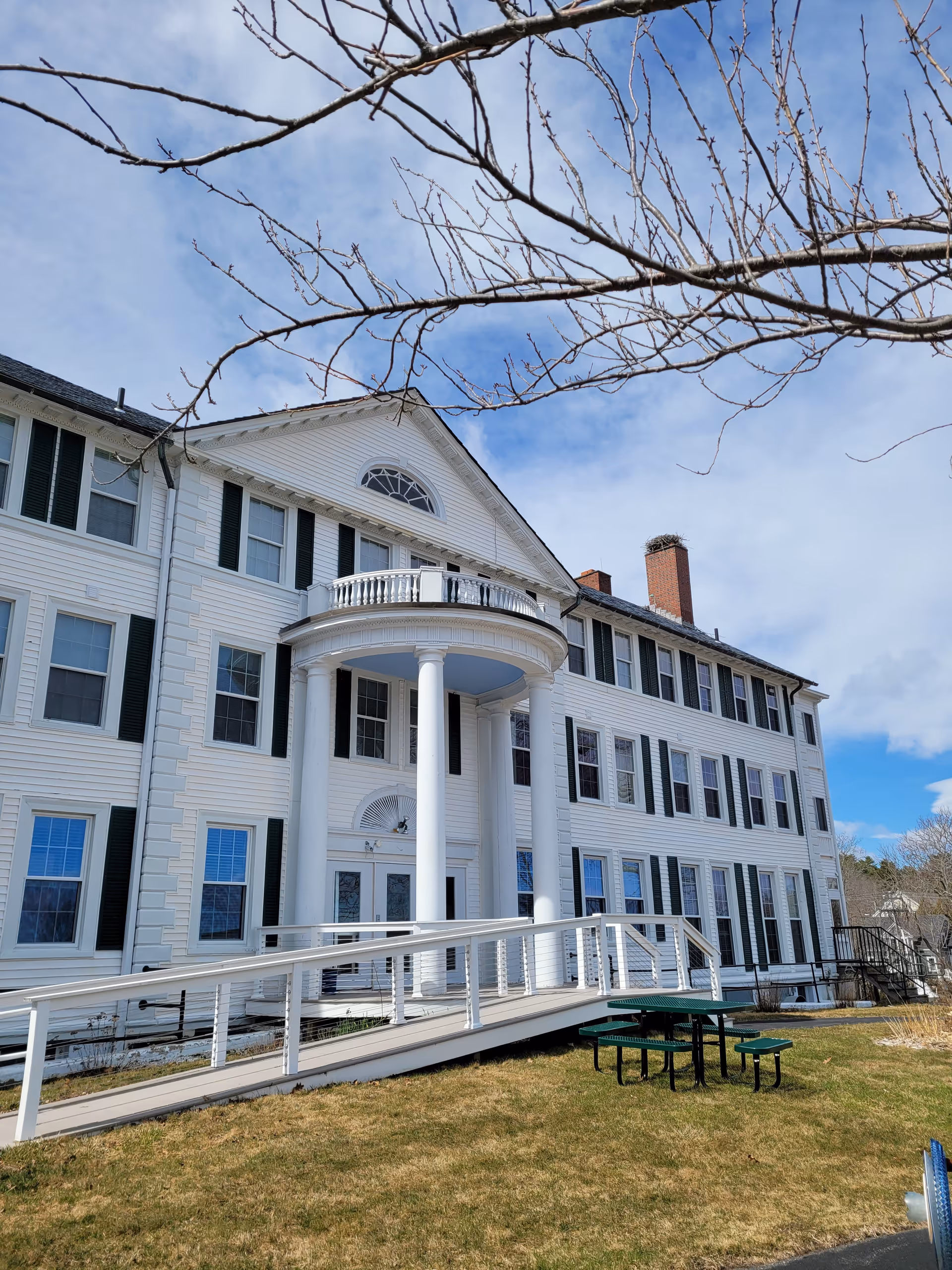 Exterior view of a large white multi-story building with black shutters and a prominent entrance featuring tall white columns and a curved balcony above. A wheelchair accessible ramp leads to the entrance. In the foreground, there is a green picnic table on a grassy area. Bare tree branches extend into the top of the image under a partly cloudy sky.