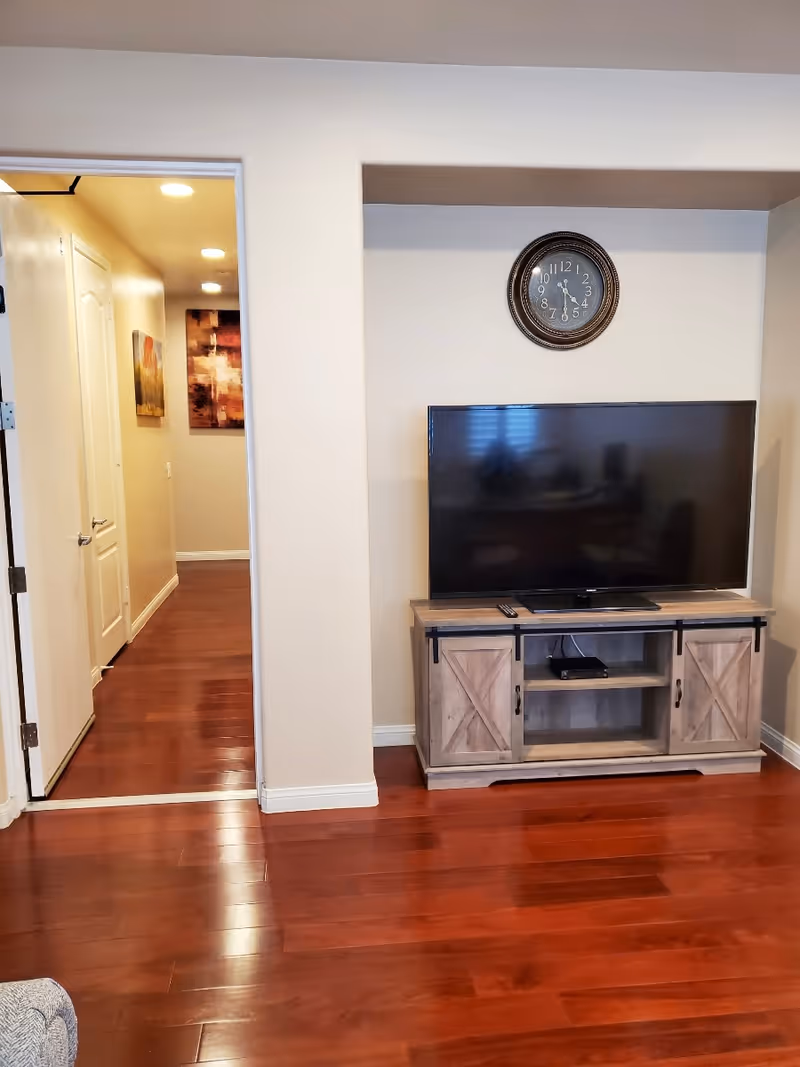 Interior view of a living room area with polished wooden floors, a flat-screen TV on a wooden TV stand, and a round wall clock above the TV. To the left, there is an open doorway leading to a hallway with wall art and recessed ceiling lights.