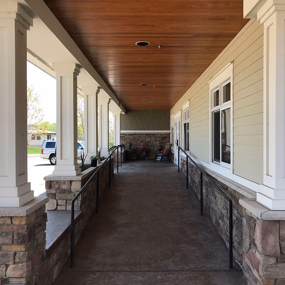 Covered front porch walkway with stone columns, handrails, and a wooden ceiling leading to the building entrance.
