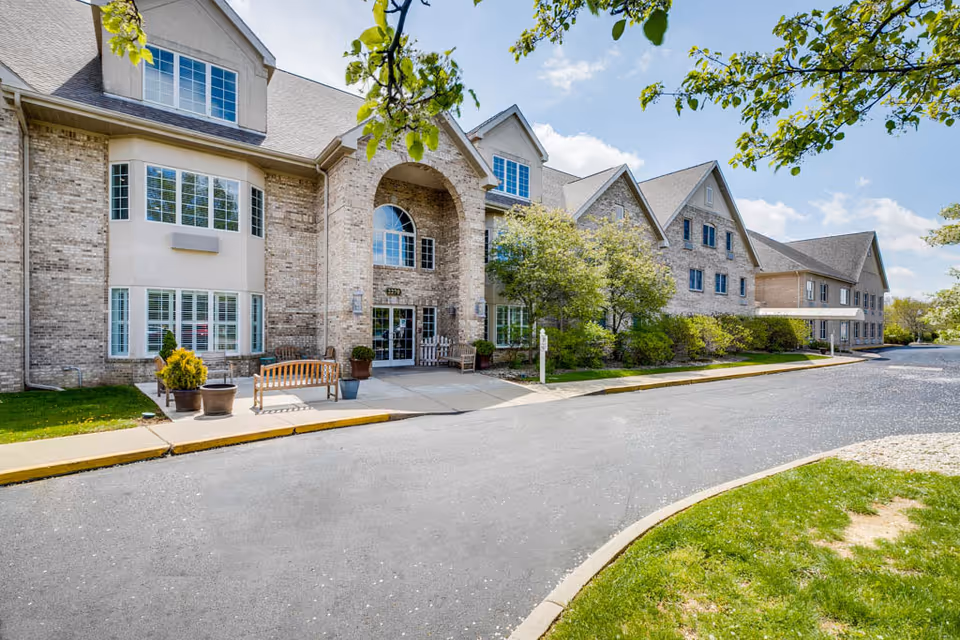 The front entrance of a brick multi-story assisted living building with benches, potted plants, trees, and a driveway.