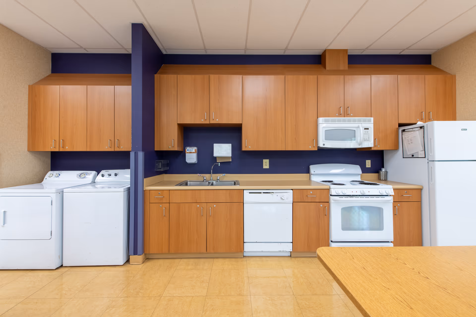 A kitchen area with wooden cabinets, a white microwave above a white stove, a white refrigerator, a dishwasher, and a double sink. To the left of the kitchen appliances, there is a white washing machine and dryer. The walls behind the cabinets are painted dark blue, and the floor is a light tan color.