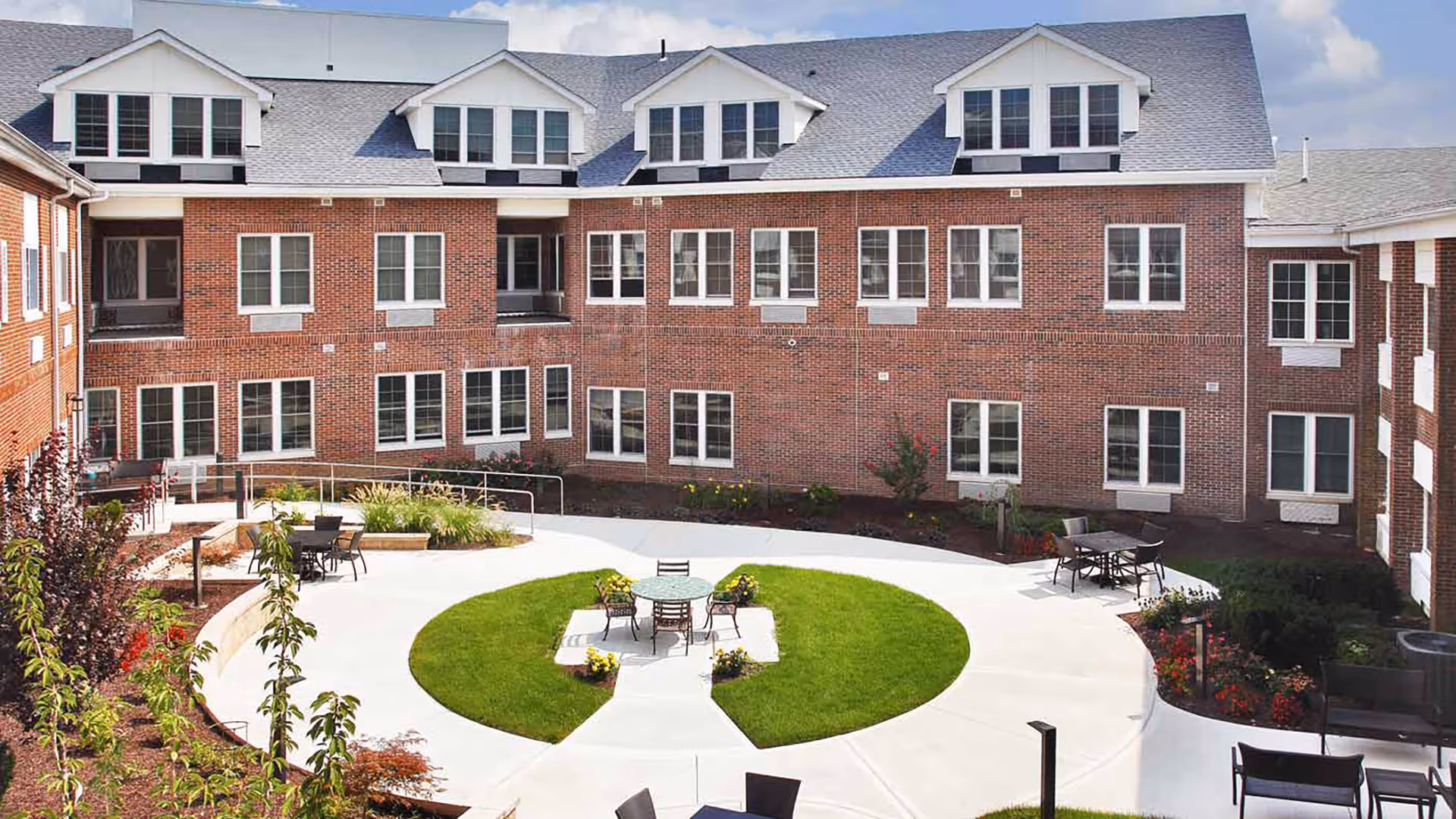 Outdoor courtyard area of a senior living facility with a circular green lawn in the center, surrounded by a concrete walkway. Several tables and chairs are placed around the courtyard, with a red brick building featuring multiple windows in the background under a partly cloudy sky.