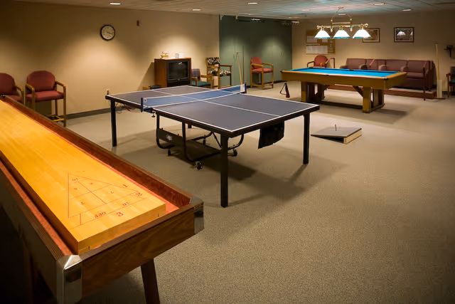 A recreational room with a shuffleboard table in the foreground, a ping pong table in the center, and a pool table in the background. The room has beige walls and carpeted floor, with several chairs and a small TV on a stand against the wall. There is a clock on the wall and framed pictures hanging above the pool table.