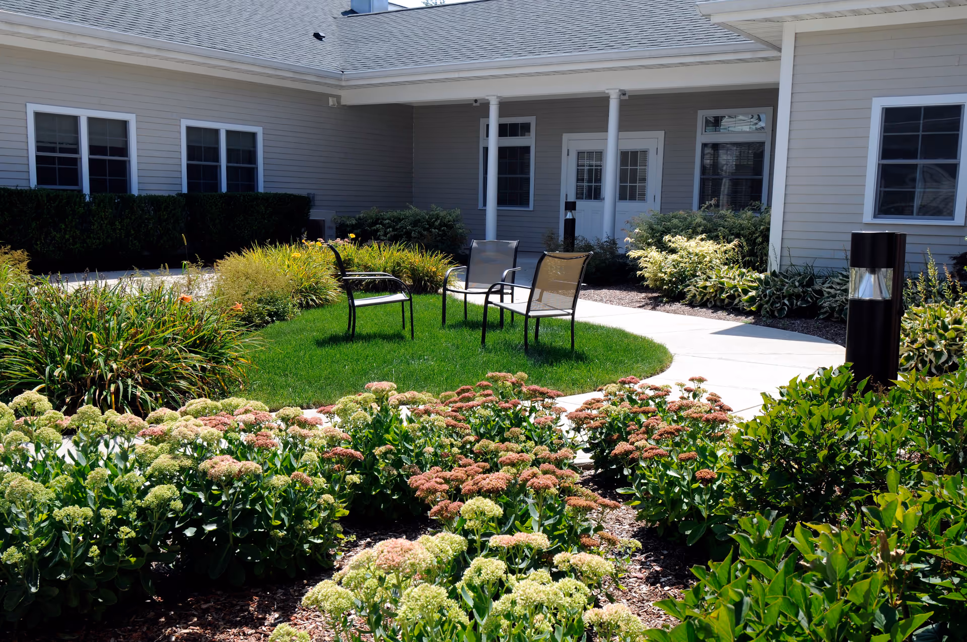 Outdoor courtyard area with green grass, flowering plants, and shrubs surrounding a circular paved walkway. Three metal chairs are placed on the grass in the center. The building exterior is light-colored with multiple windows and a door leading inside.