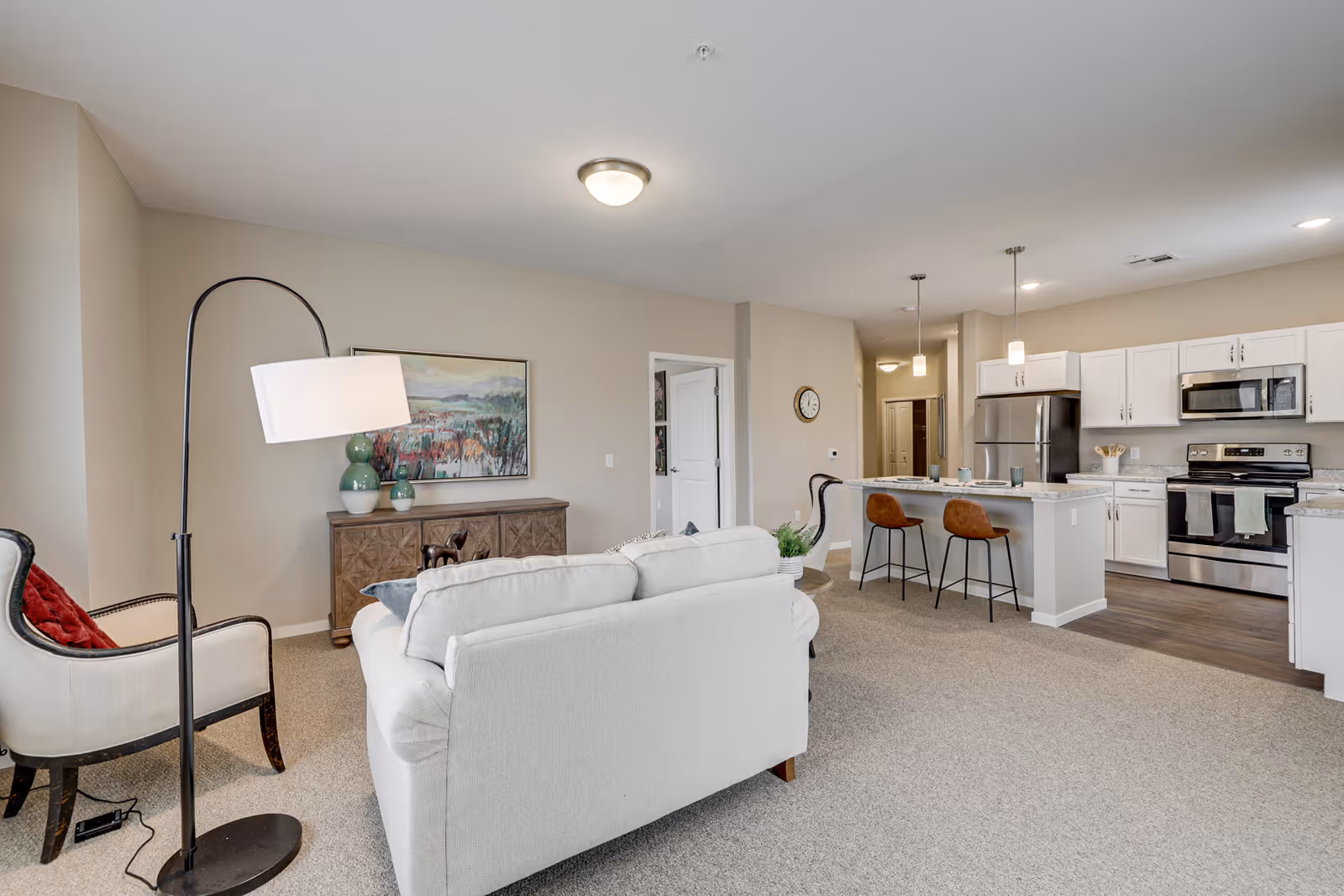 A bright and spacious open-concept living area in a senior living facility featuring a white sofa, an armchair with a red cushion, a floor lamp, a wooden sideboard with decorative vases and a landscape painting above it. The living area opens into a modern kitchen with white cabinets, stainless steel appliances, a kitchen island with two brown bar stools, and pendant lighting.