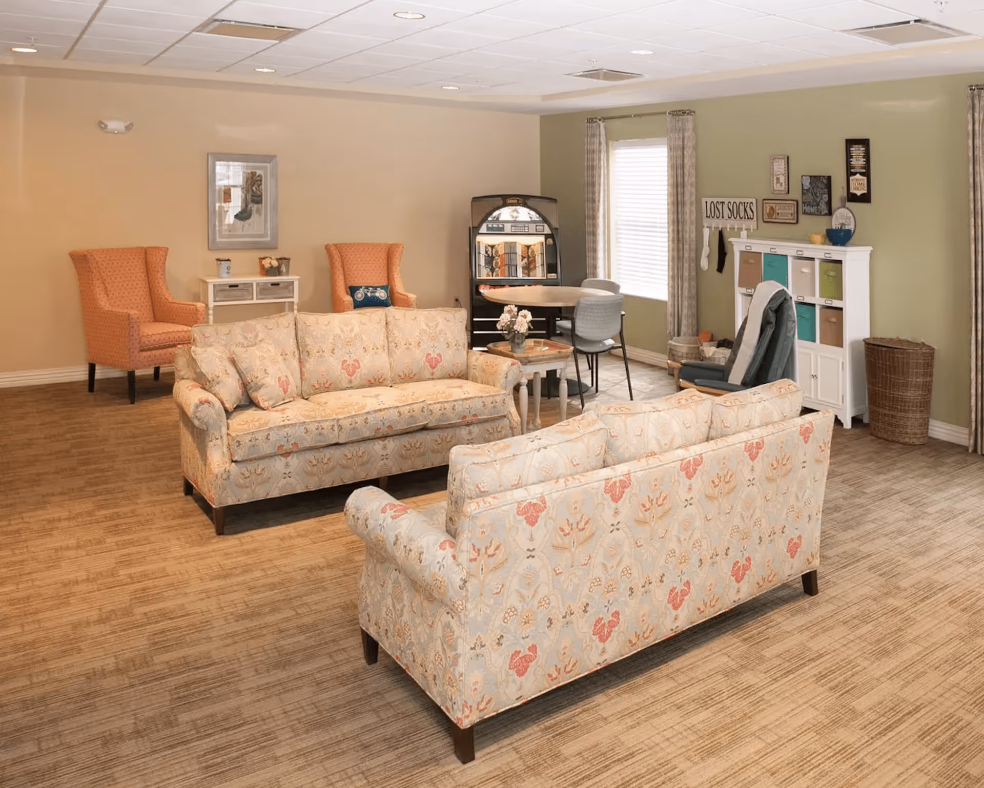 A cozy living room area with two patterned sofas facing each other, two orange armchairs in the background, a small table with flowers, a round table with chairs near a window, and a white storage cabinet with colorful bins and wall decorations including a 'LOST SOCKS' sign.