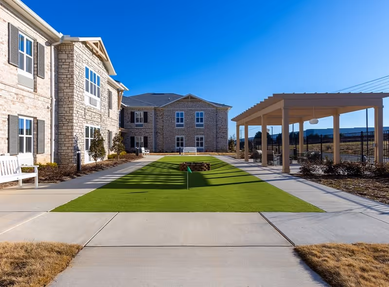 Courtyard of a senior living facility with a central putting green, stone buildings, benches, and a covered pergola under a clear blue sky.